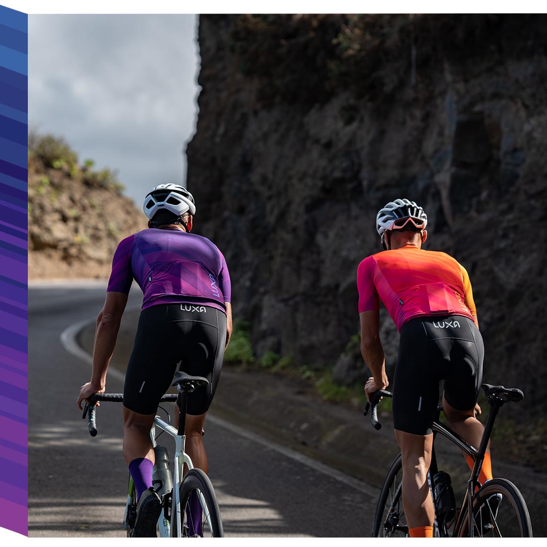 Two male cyclists in Luxa cycling outfits - one in a purple jersey and shorts, the other in an orange jersey and shorts. One of the cyclists is wearing orange socks to match the color of his jersey.